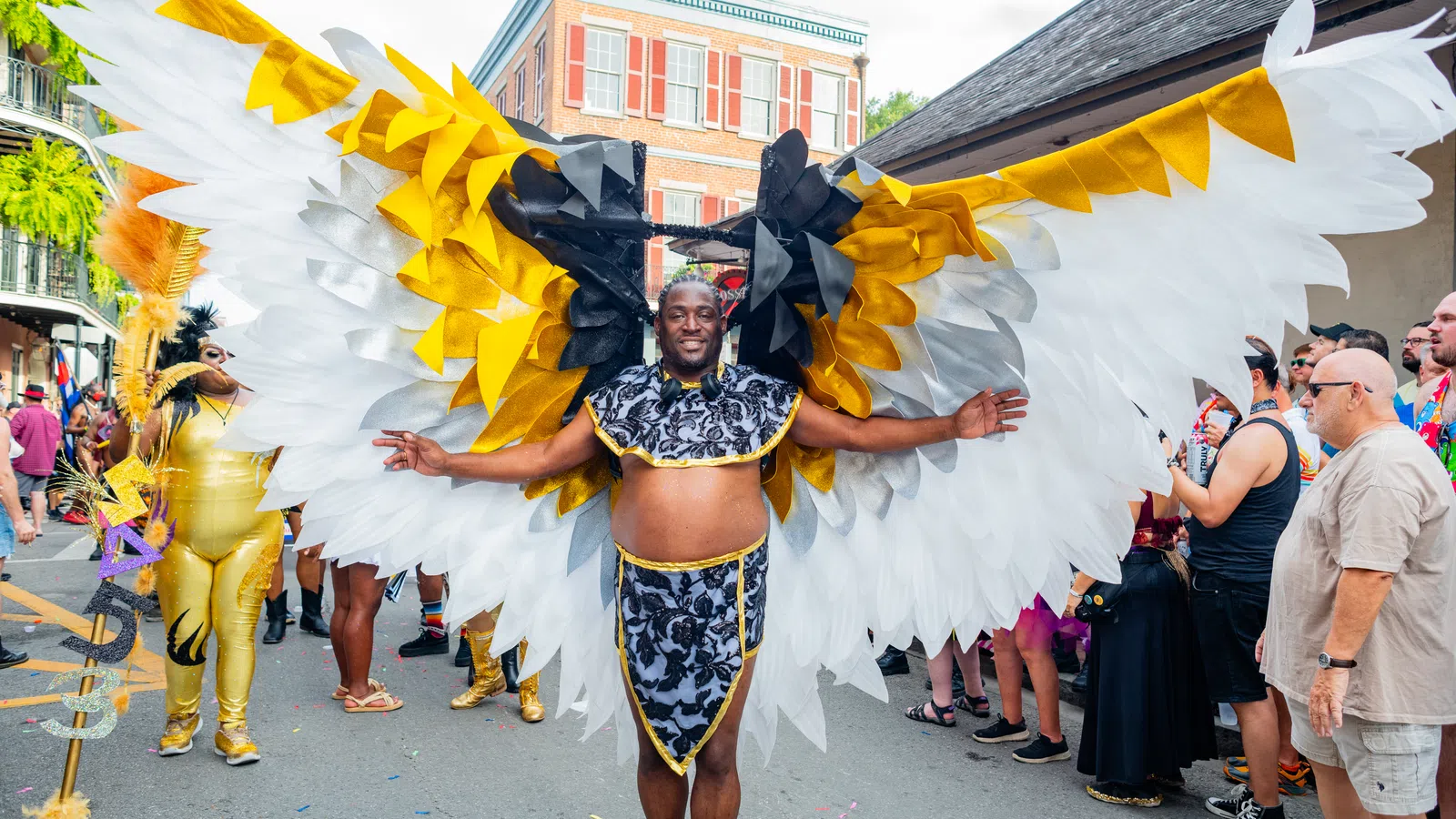 Man wearing large white, gold, and black wings costume at colorful outdoor street festival parade.