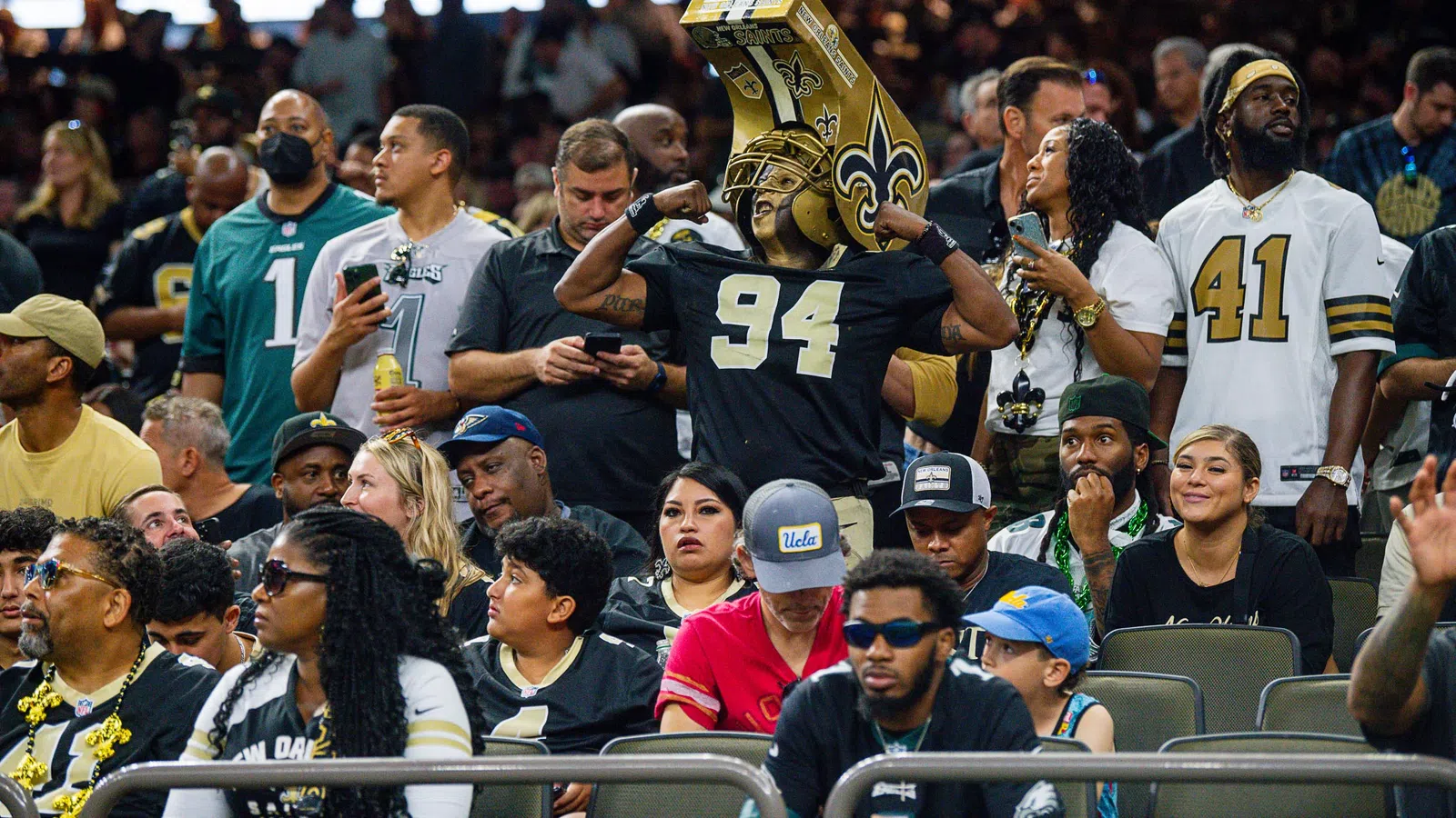 Excited fans at football game, some wearing New Orleans Saints jerseys and merchandise in stadium seats.