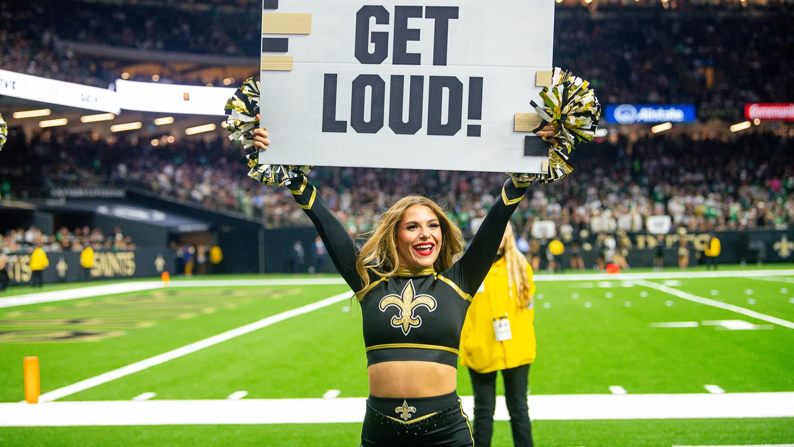 Cheerleader in New Orleans Saints uniform holding a GET LOUD! sign on football field with crowd behind