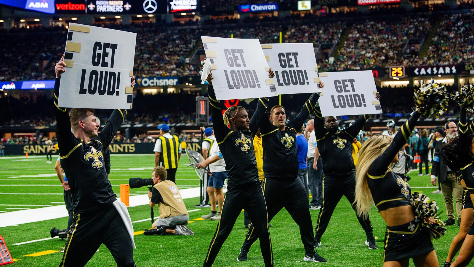 New Orleans Saints cheerleaders and dancers holding Get Loud signs on the football field during a game.