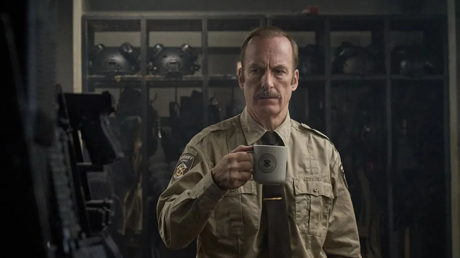 Police officer in uniform holding a white mug inside an armory with helmets and gear in the background