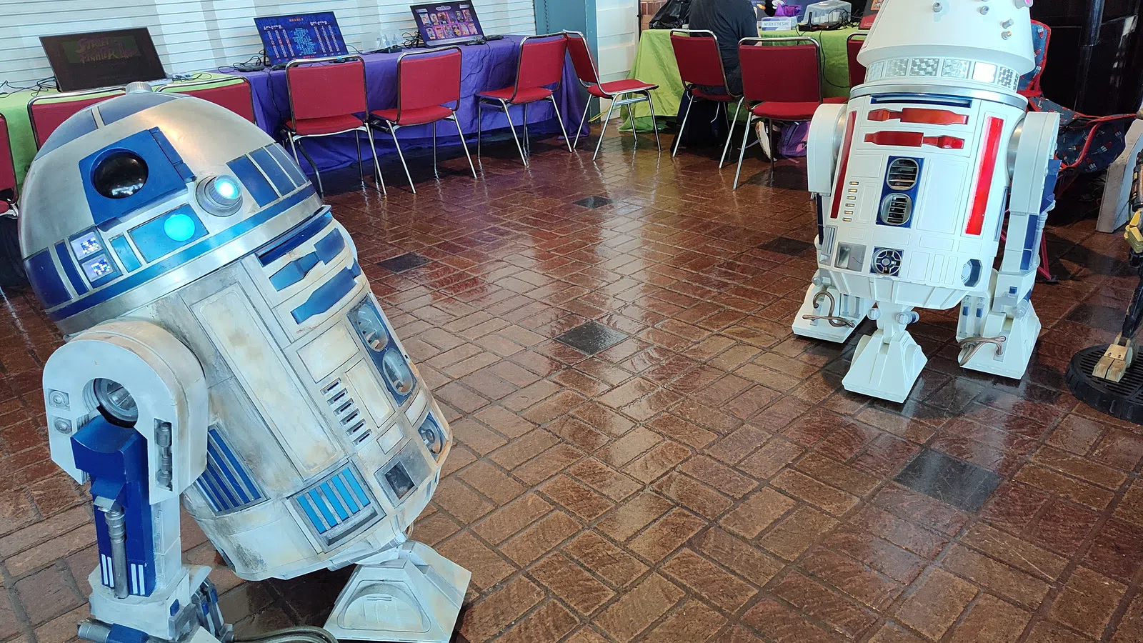 Two detailed Star Wars droids R2-D2 and an R2 variant stand on a tiled floor in a convention setting with laptops and chairs.