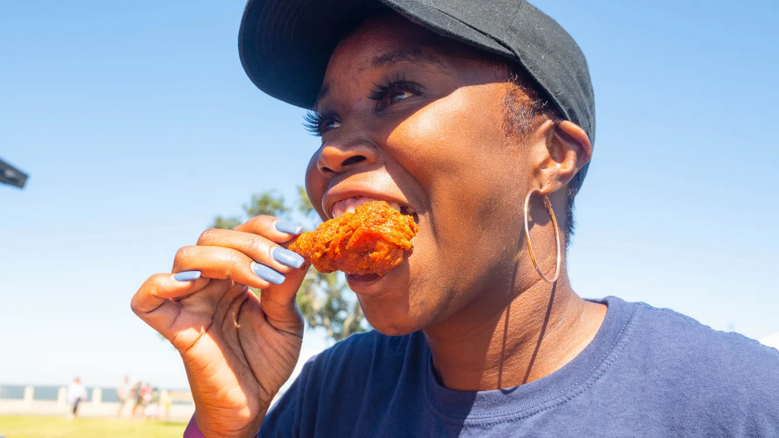 Smiling woman wearing a black cap and hoop earrings eating fried chicken outdoors on a sunny day