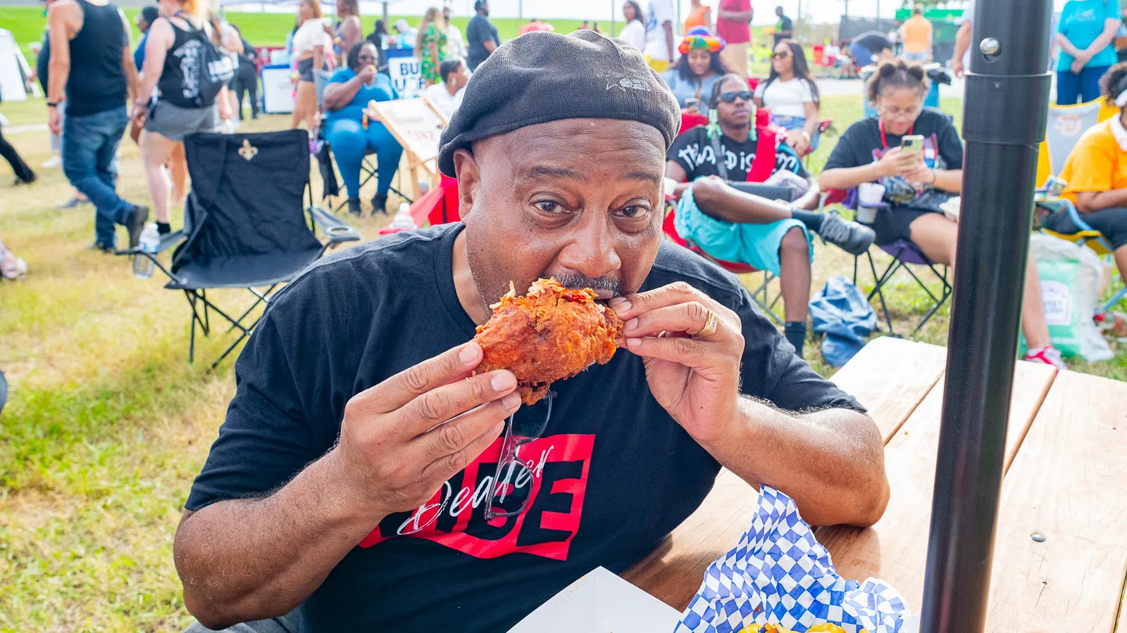 Man in black hat eating fried chicken at outdoor event with people and tents in the background