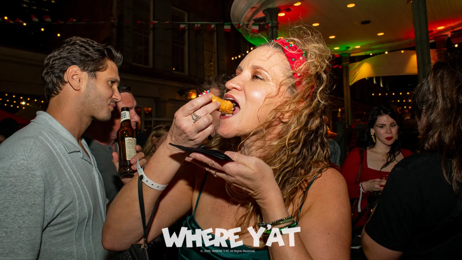 Woman in red headband eating fried food at lively nighttime party with people drinking and socializing.