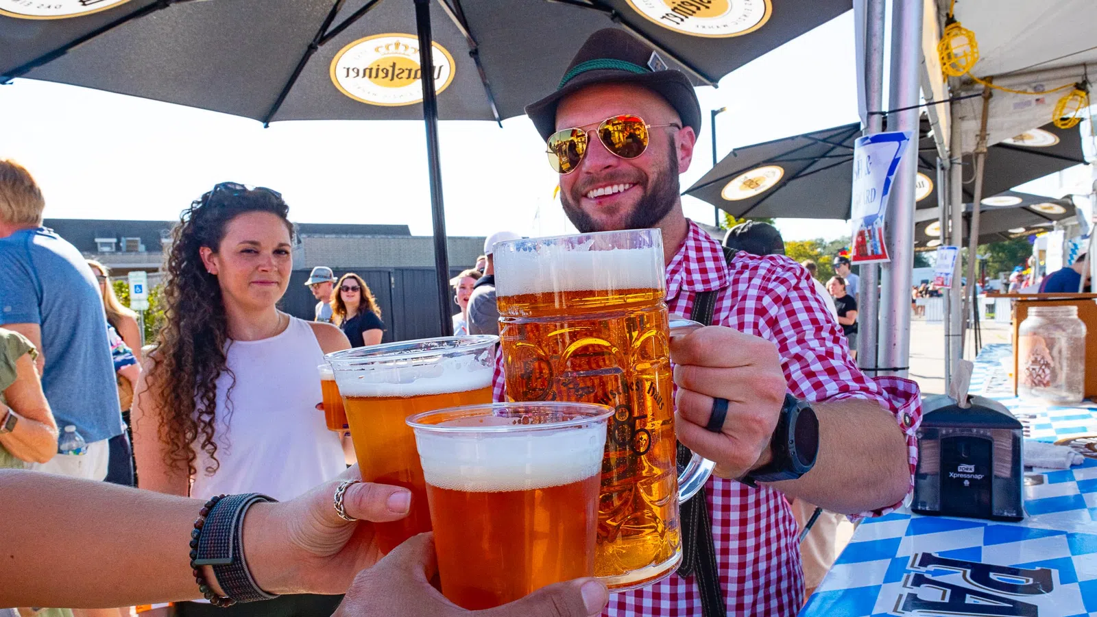 Group of people clinking large beer mugs and cups at an outdoor festival under branded umbrellas