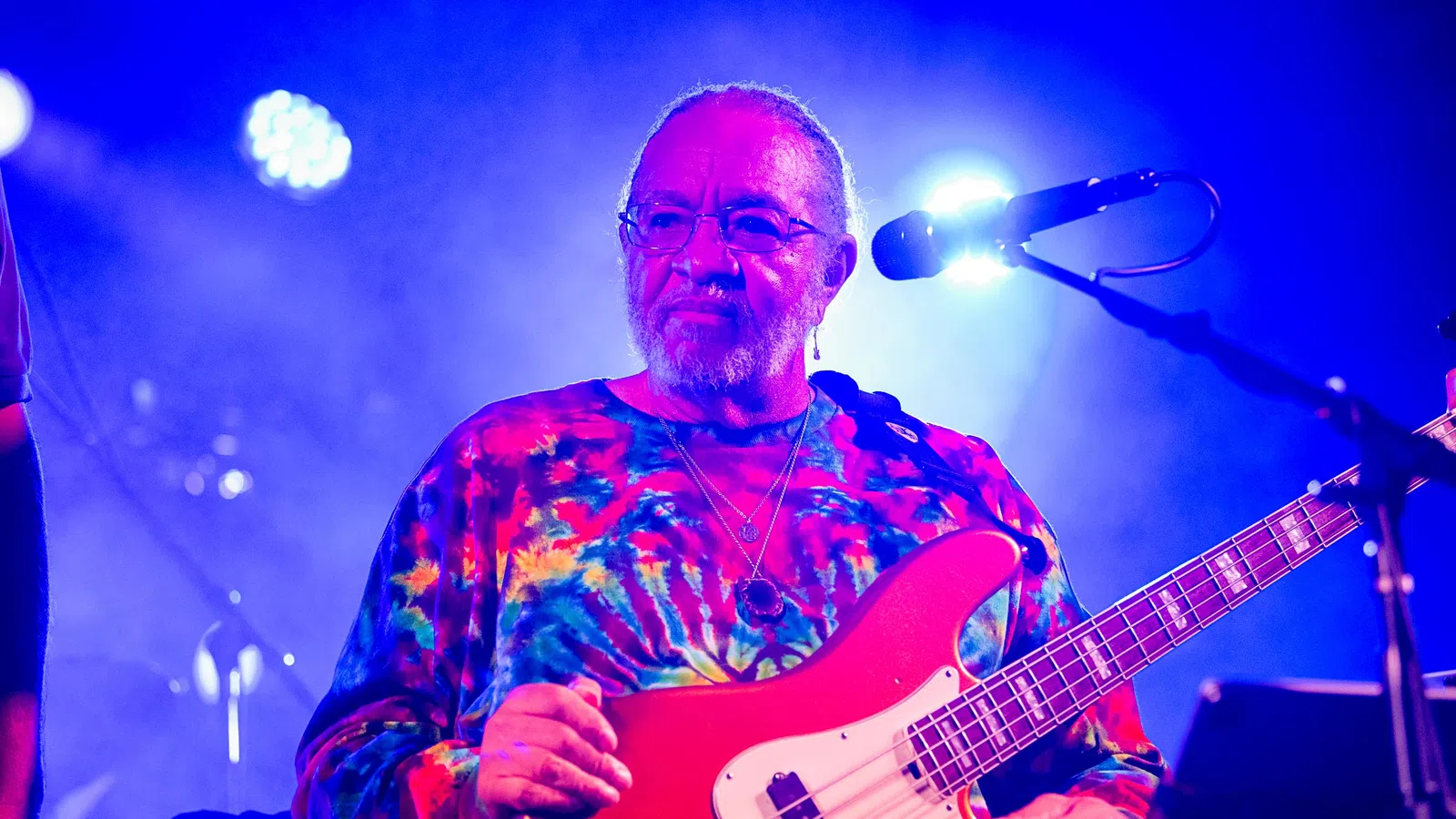 Musician wearing a colorful tie-dye shirt playing red electric bass guitar on stage with blue lighting.