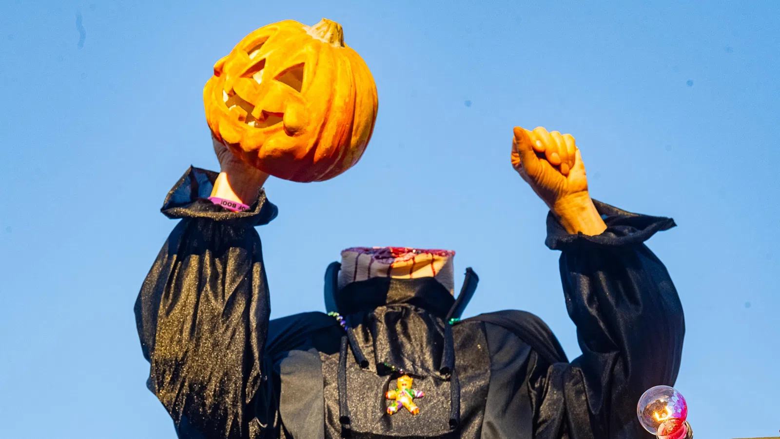 Headless Halloween figure in black costume holding a carved pumpkin against a clear blue sky.