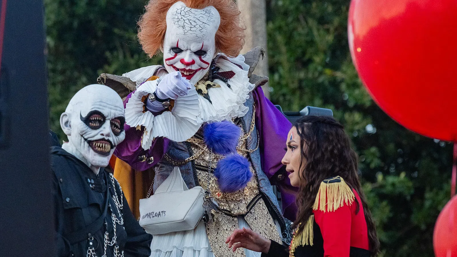 Three people in horror costumes including a clown with red hair, a skull-faced figure, and a woman in a red and gold jacket.