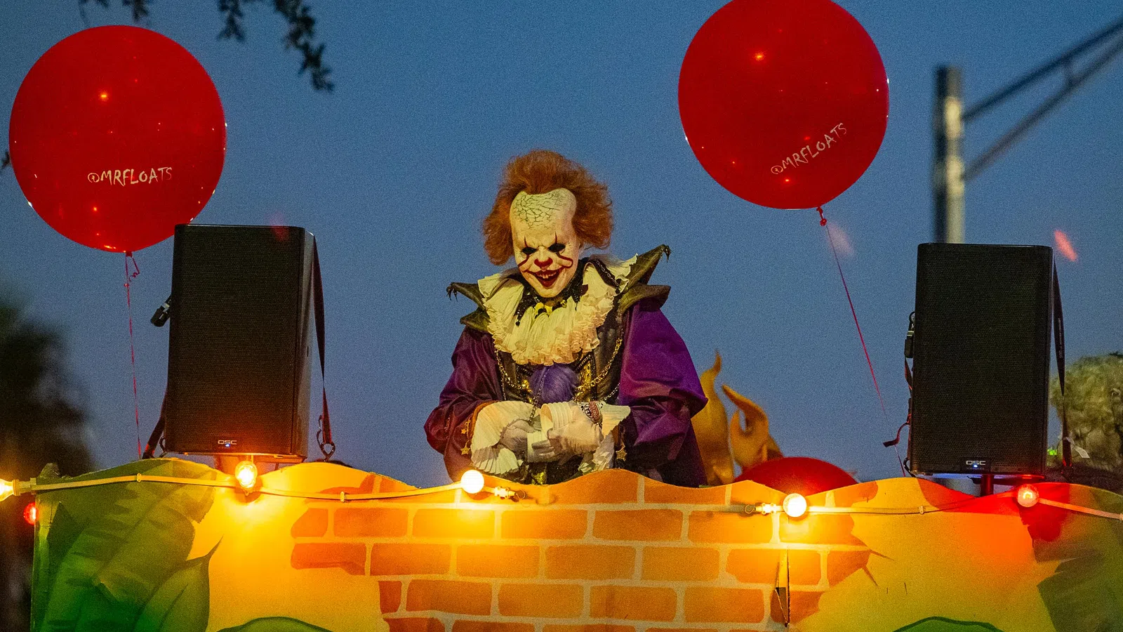 Costumed clown with red hair and white face paint holding red balloons at night during parade float event.