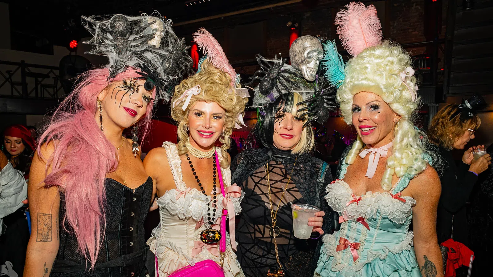 Four women in elaborate Halloween costumes with wigs and accessories at a festive party indoors.
