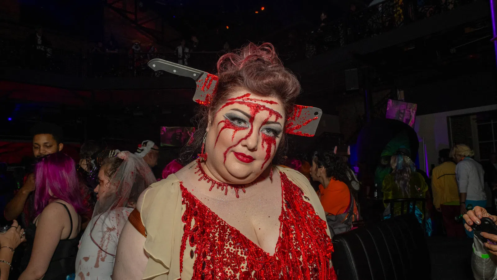 Woman in bloody costume with fake cleaver through head at a Halloween party with crowd in background