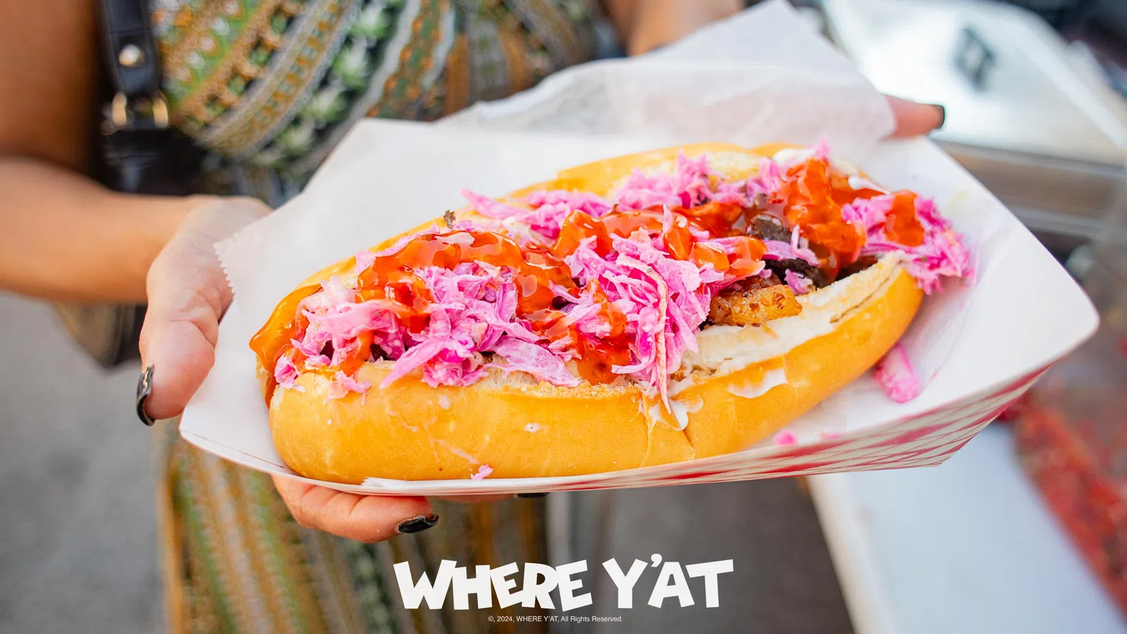 Close-up of a hand holding a hot dog with pink slaw and red sauce in a paper tray, with overlaid text reading WHERE Y'AT.