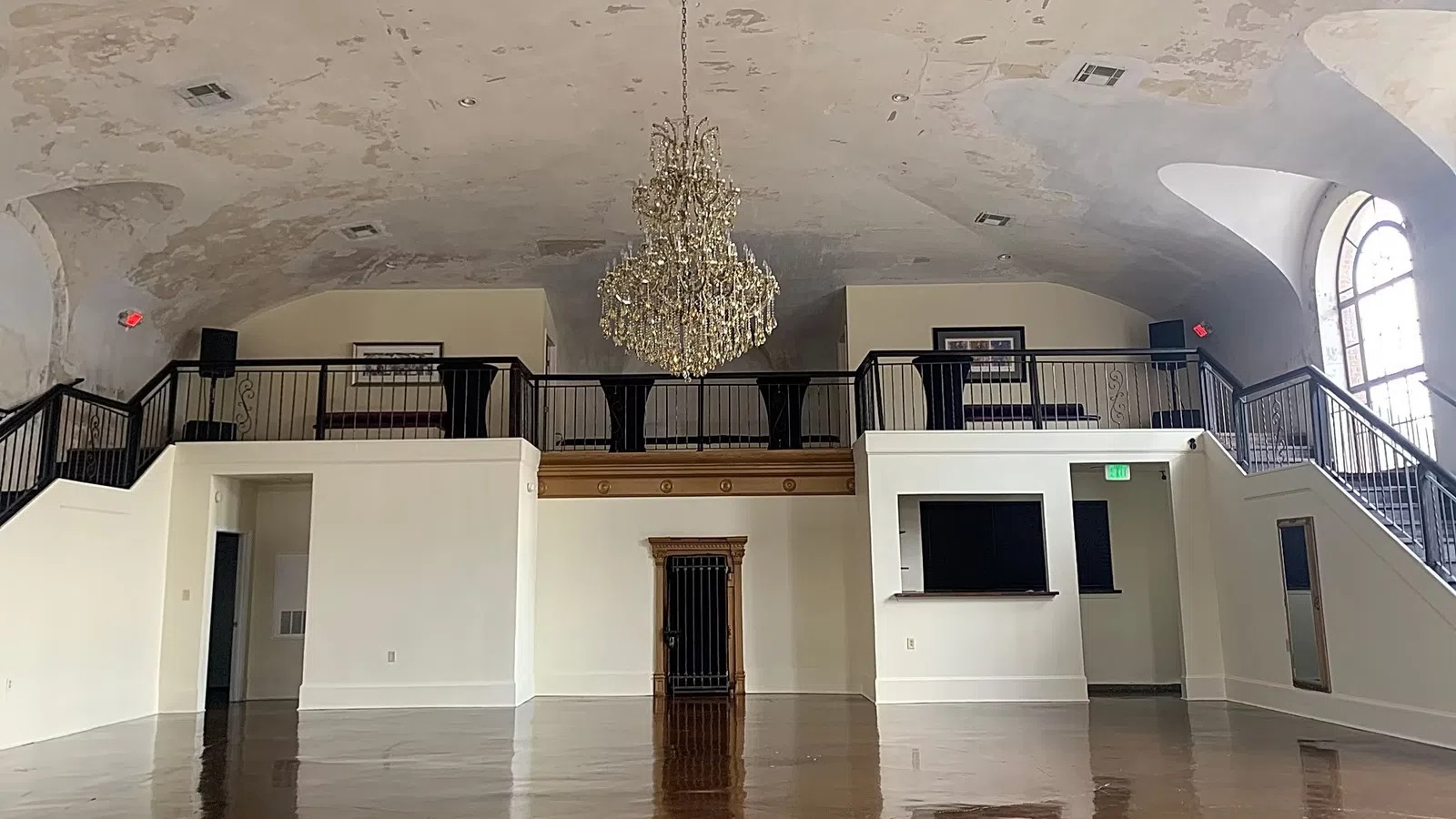 Empty spacious room with polished floor, decorative chandelier, balcony with railings, and peeling ceiling paint.