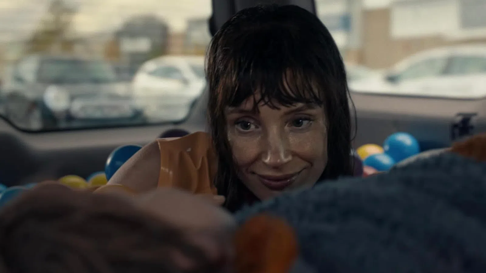 Woman with wet hair smiling inside a car filled with colorful plastic balls, looking at someone out of focus.