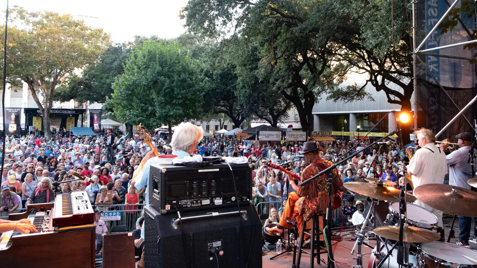View from stage of a live band performing to a large outdoor crowd under trees at a music festival.