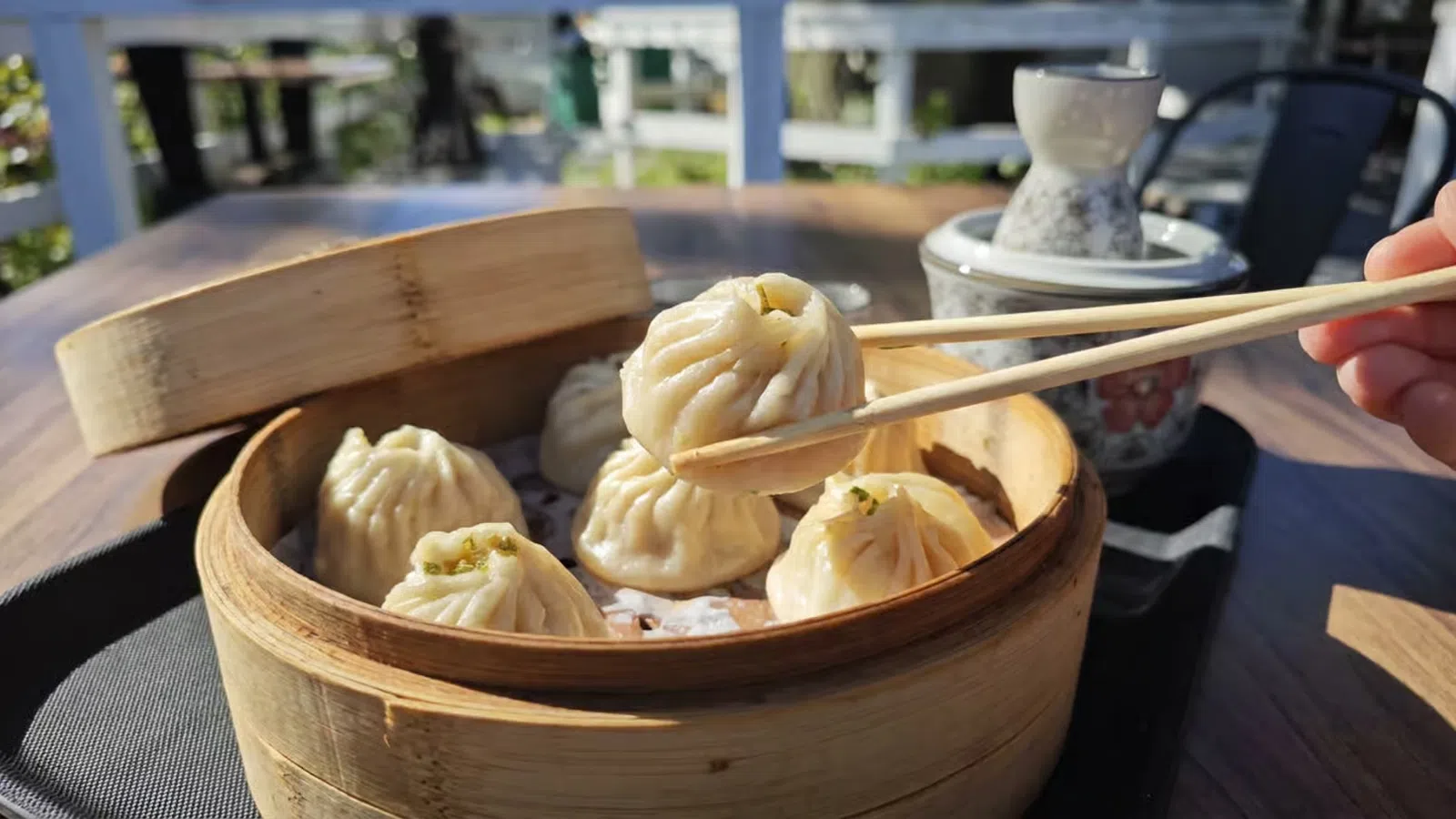 Chopsticks picking up a steamed dumpling from a bamboo basket on a wooden table outdoors