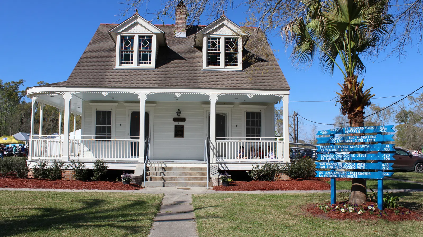 White historic house with a wraparound porch, manicured lawn, and a blue directional sign near a palm tree.