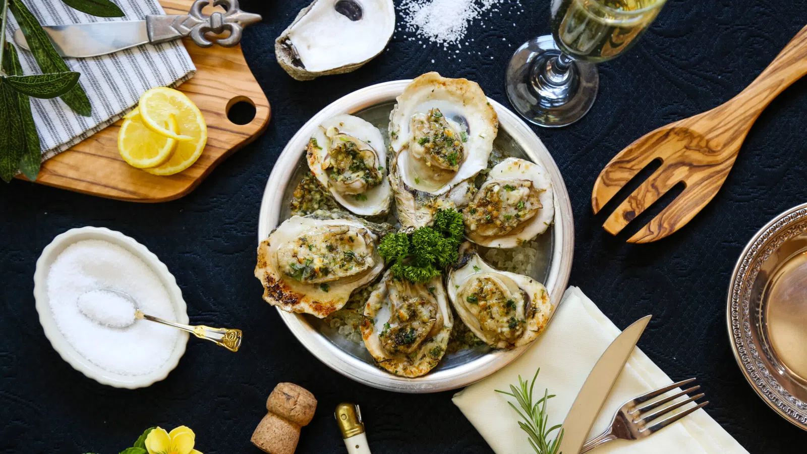 Grilled oysters on plate with lemon, salt, herbs, knife, fork, and glass of white wine on black table.