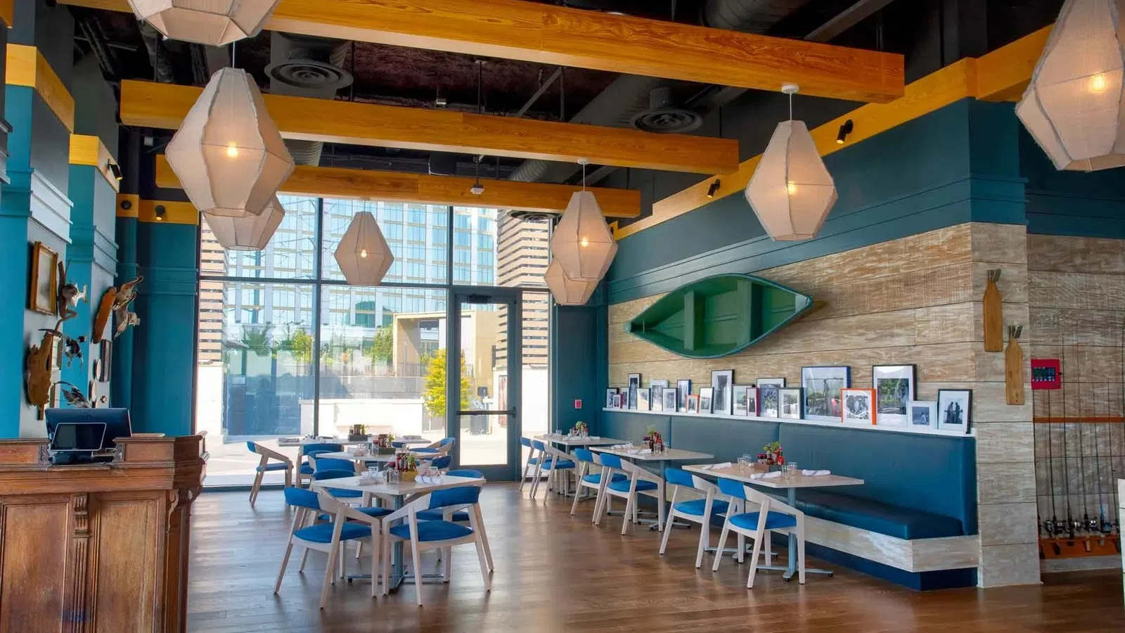 Modern dining area with wooden beams, blue chairs, pendant lights, and wall-mounted boat decor in a bright restaurant.
