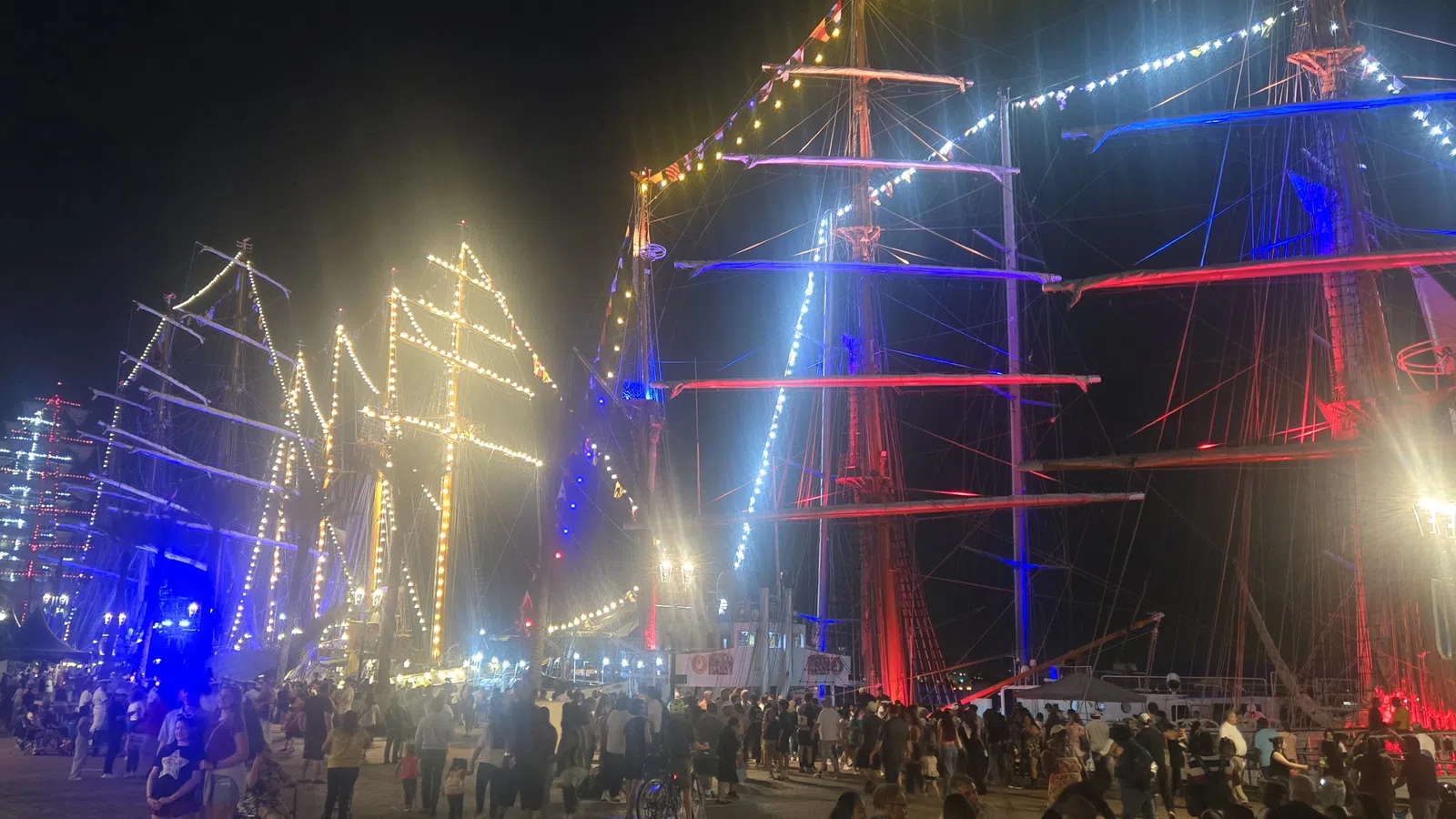 Crowd gathers at night by tall ships decorated with colorful lights and illuminated sails at a festive event.