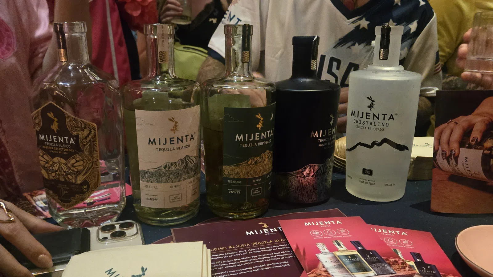 Variety of Mijenta tequila bottles displayed on table with promotional cards and people in the background.