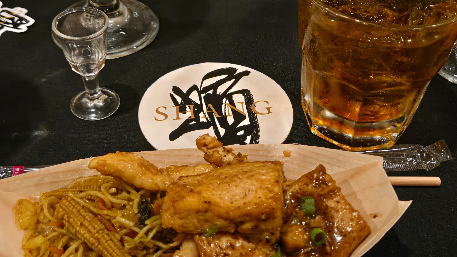 Asian stir-fry with grilled chicken, baby corn, noodles, and iced whiskey on a black table with shot glass.