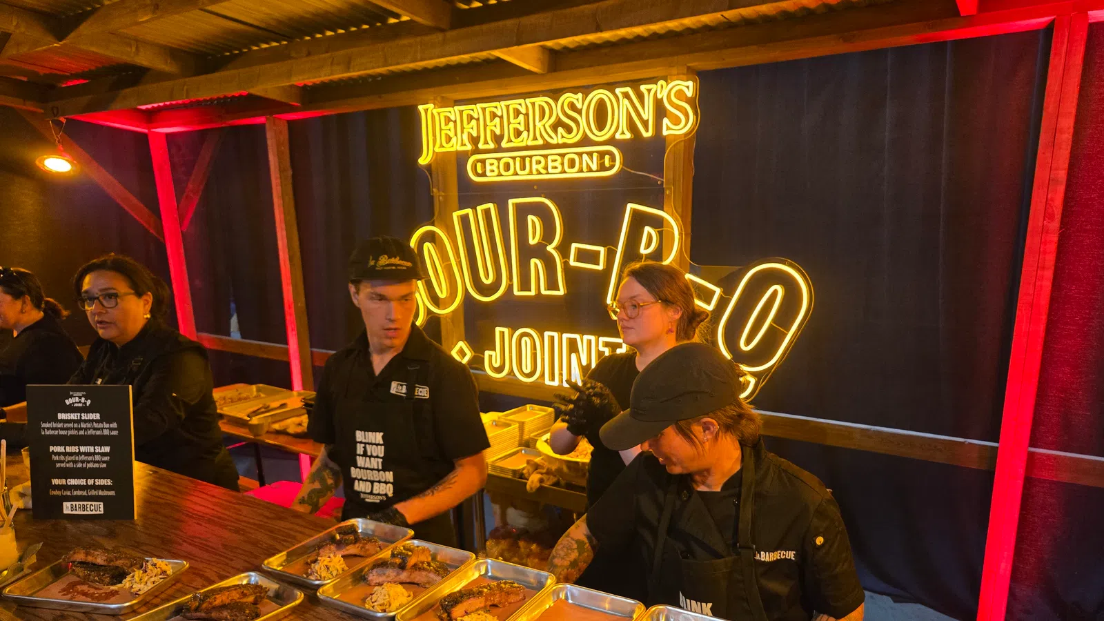 Food stall serving barbecue with brisket and ribs under bright Jefferson's Bourbon neon sign at event