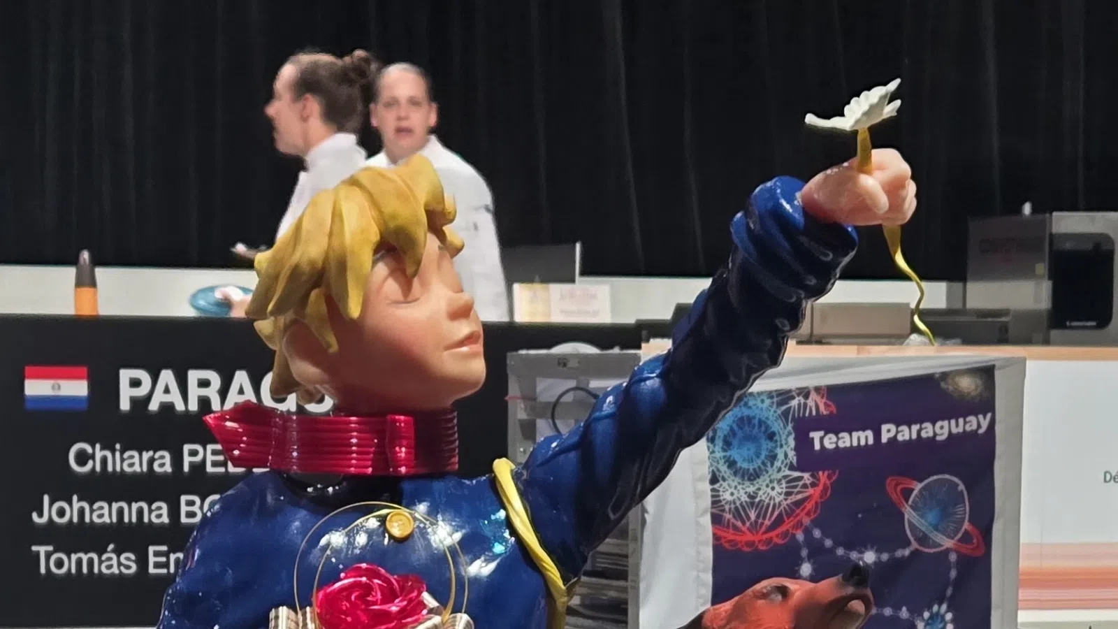 Colorful statue of a boy in blue holding a flower and a red rose with a fox looking up, Paraguay team banner in background