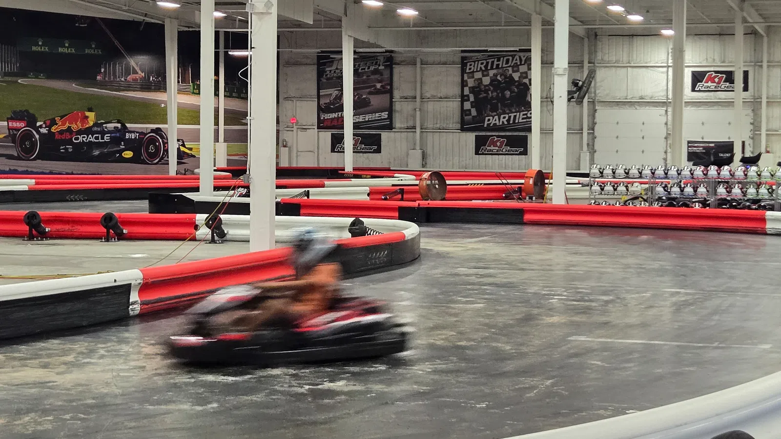 Indoor go-kart racing track with blurred driver speeding around the corner, red and white barriers, and racing posters.
