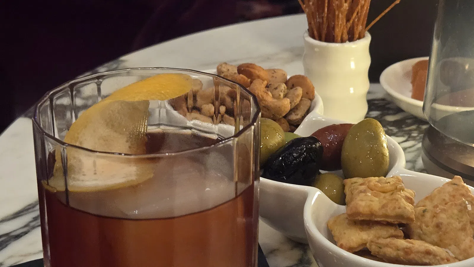 Old fashioned cocktail with lemon peel served with assorted nuts, olives, crackers, and breadsticks on marble table.