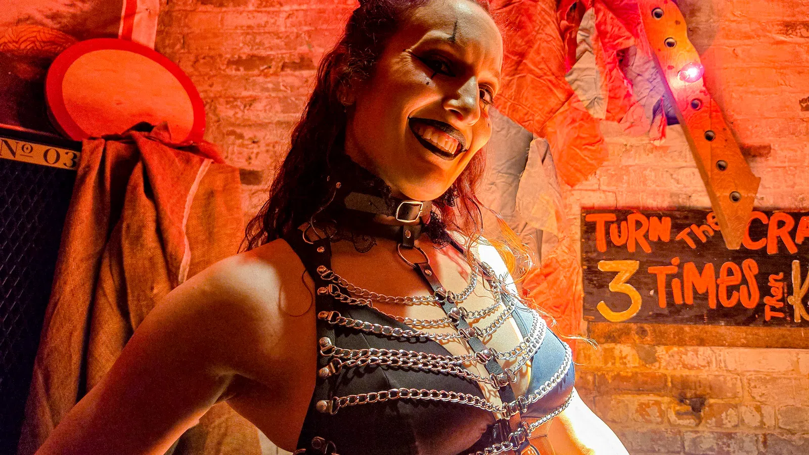 Woman in black leather harness and chains smiles against a brick wall with Halloween decor and overlaid text sign.