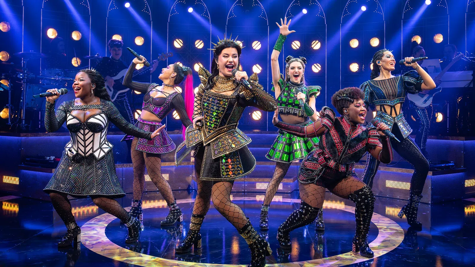 Six female performers sing and dance energetically on a brightly lit stage with colorful costumes and fishnet stockings.