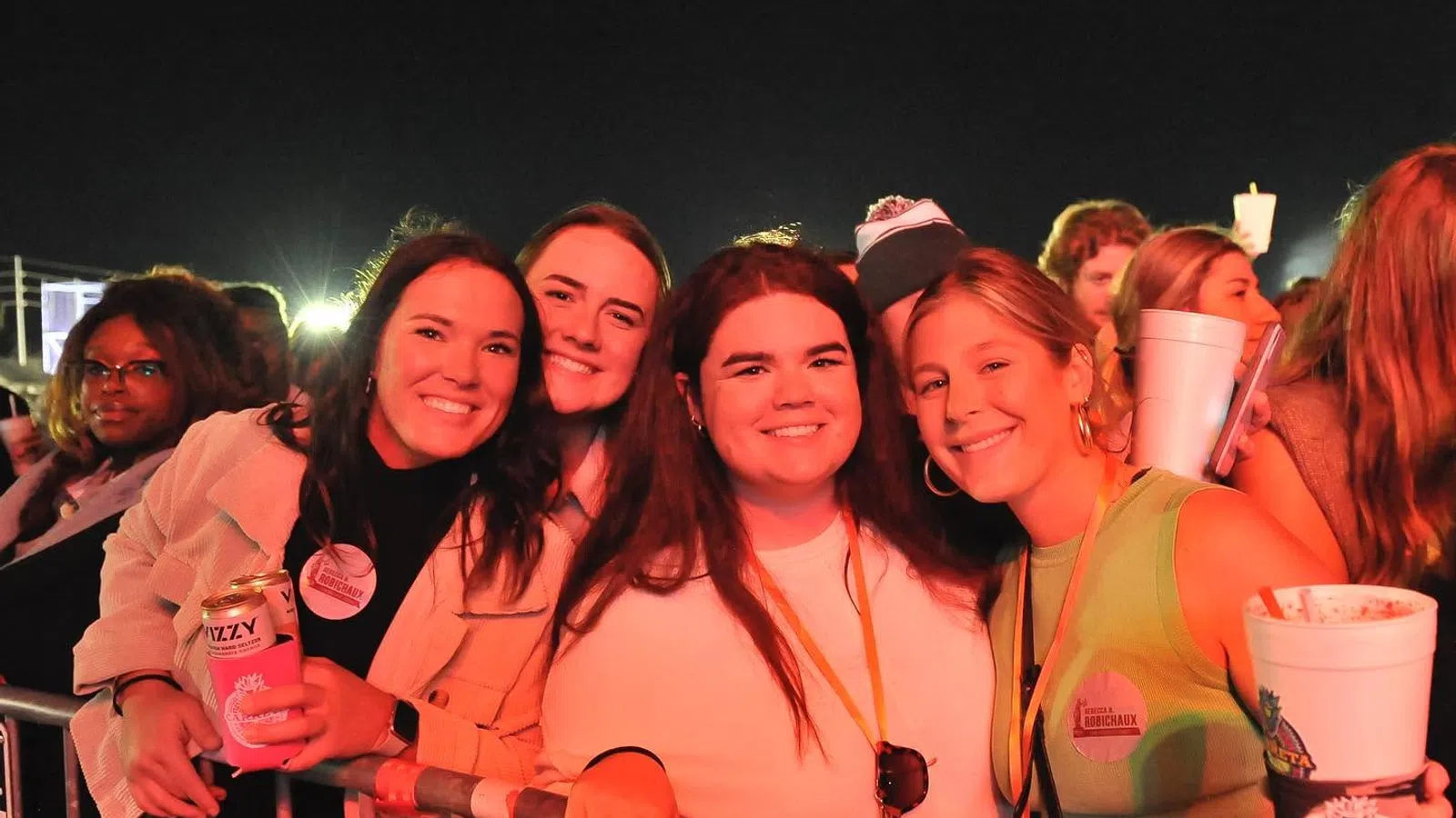 Four smiling young women posing behind a metal barrier at a nighttime outdoor event or concert, holding drinks.