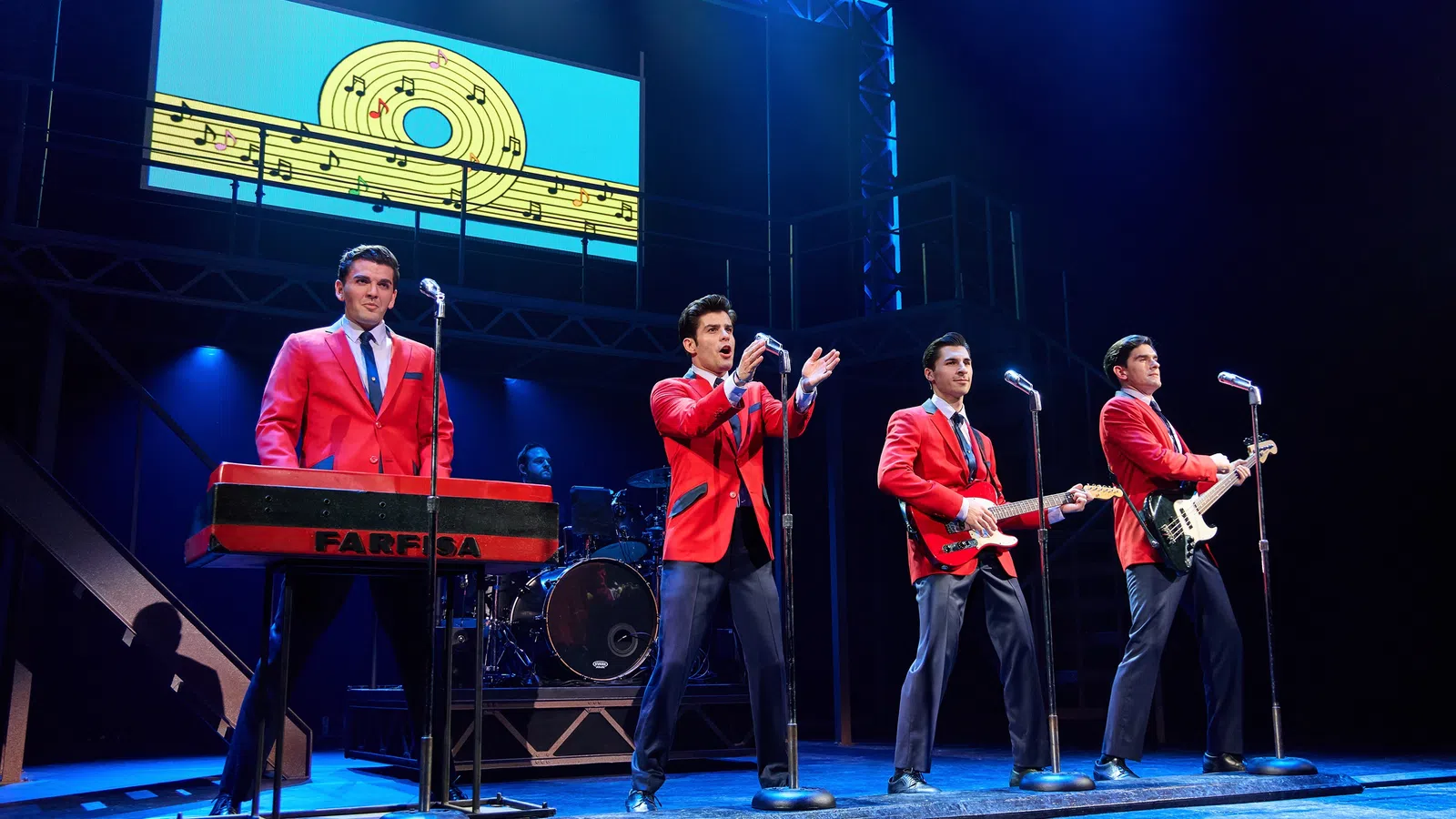 Four male performers in red jackets play instruments and sing on stage with musical notes backdrop in blue lighting.
