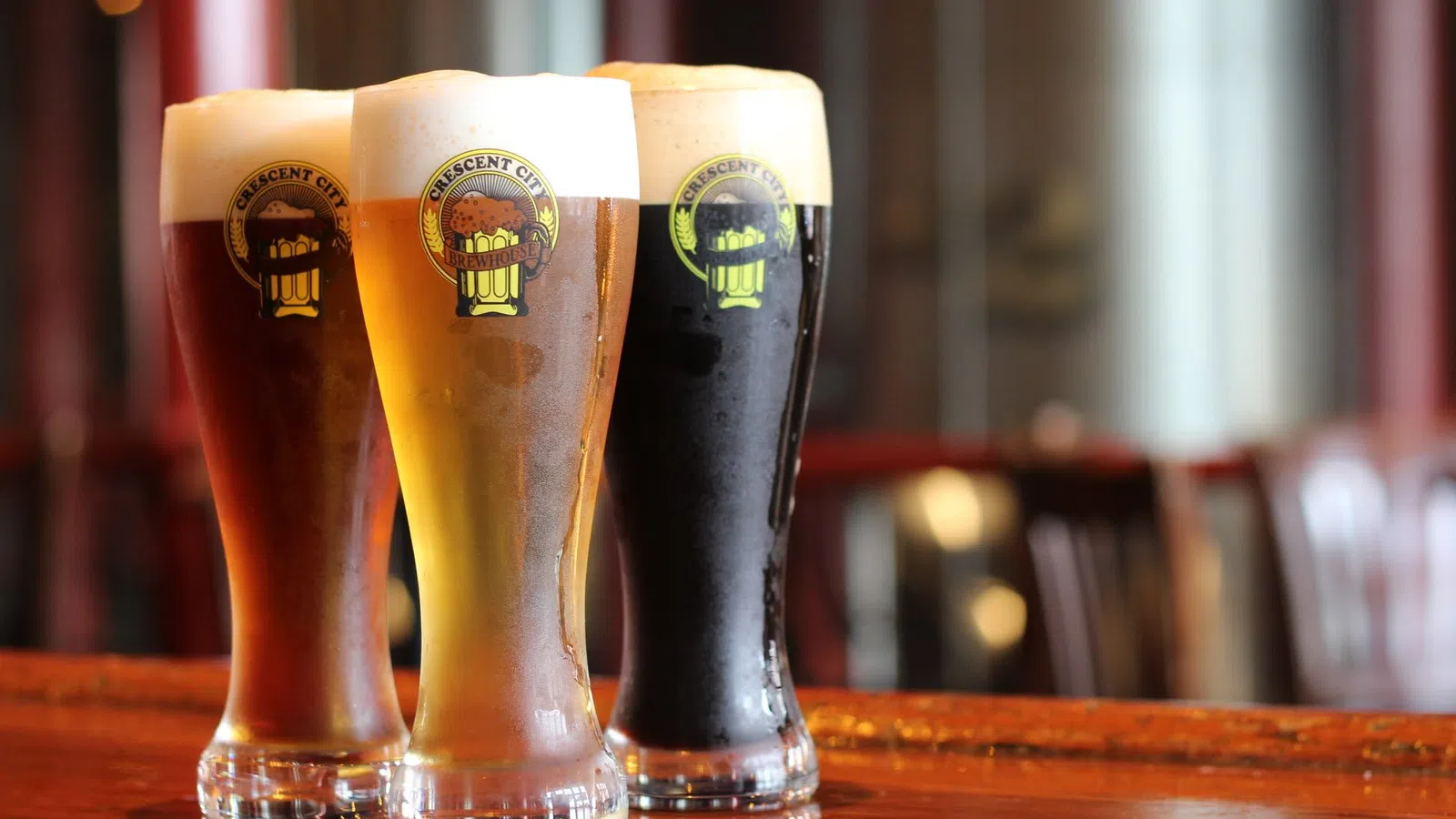 Three tall glasses of beer with varying colors and foam on a wooden bar counter in a pub setting.