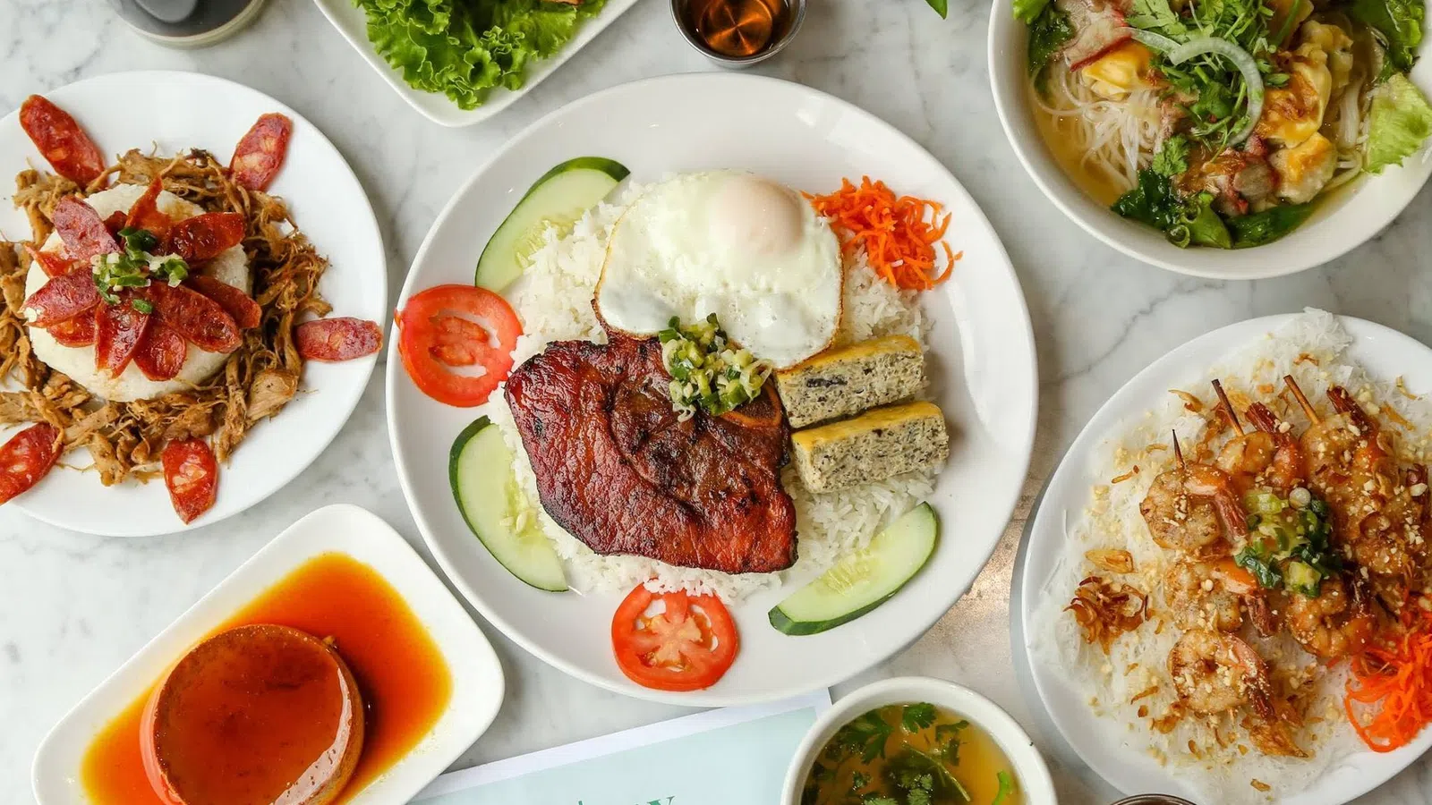 Overhead view of Vietnamese dishes including grilled pork with egg, spring rolls, noodle soup, shrimp skewers, and caramel flan on a marble table.