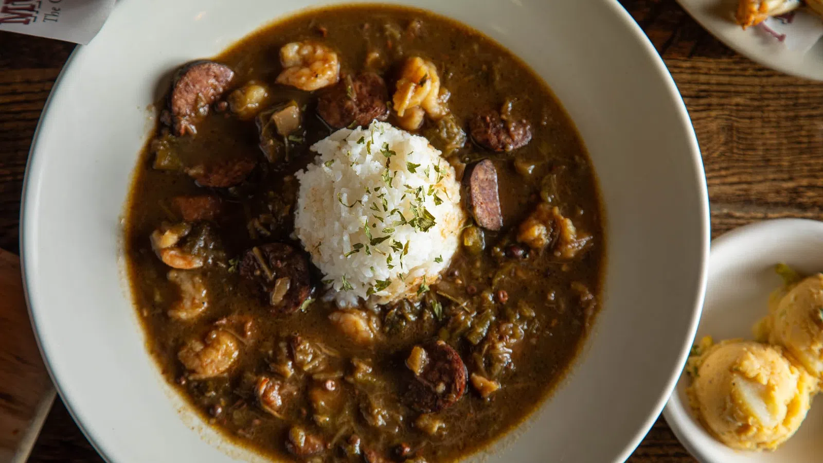 Bowl of shrimp and sausage gumbo with white rice and herbs on top, served on a wooden table with side dishes.