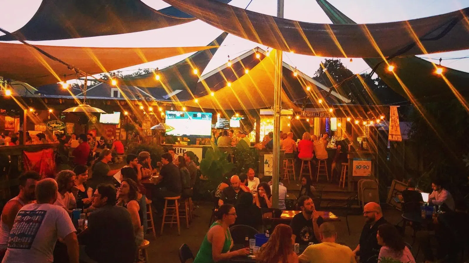 Outdoor evening bar scene with string lights, shaded canopies, and groups of people socializing under warm lighting.