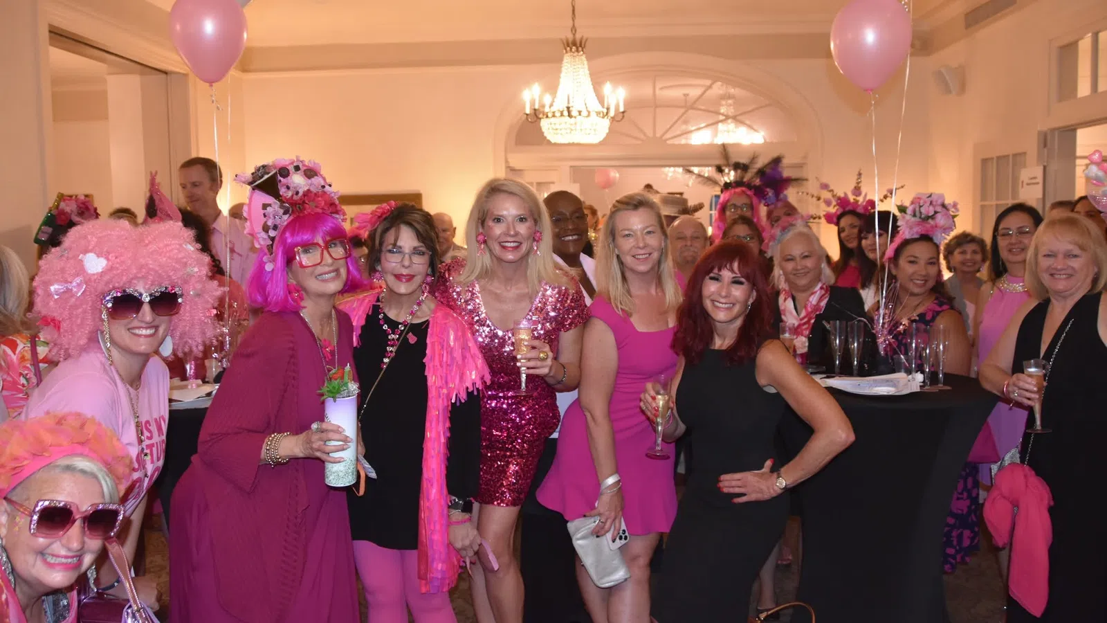 Group of women dressed in pink and black at an indoor party with balloons and drinks celebrating together.