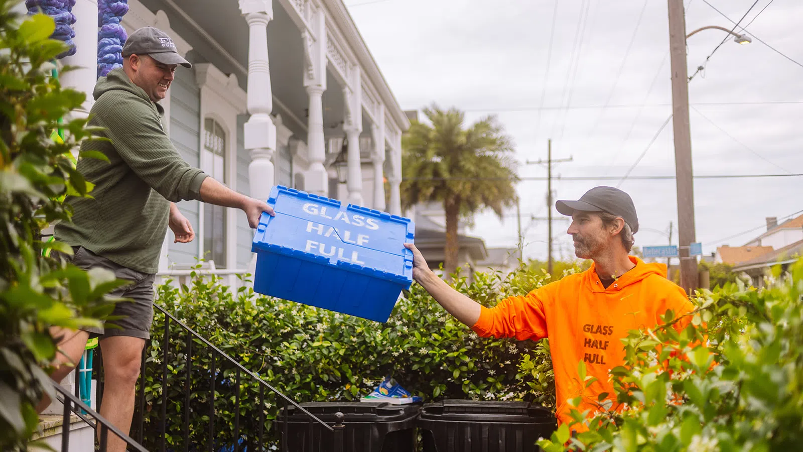 Two men exchanging a blue recycling bin labeled GLASS HALF FULL in a residential outdoor setting with greenery.