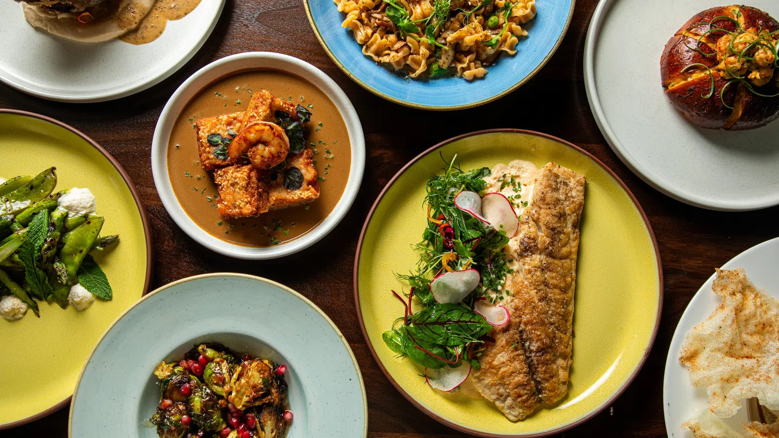 Overhead view of diverse gourmet dishes including fish filet, pasta, roasted vegetables, and appetizers on colorful plates.