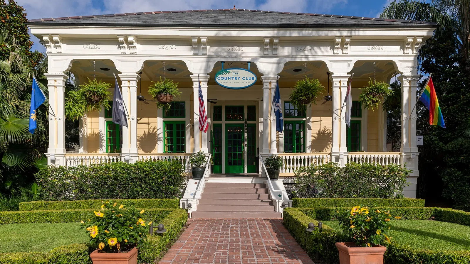 Historic country club entrance with hanging plants, flags, brick walkway, and manicured garden under blue sky