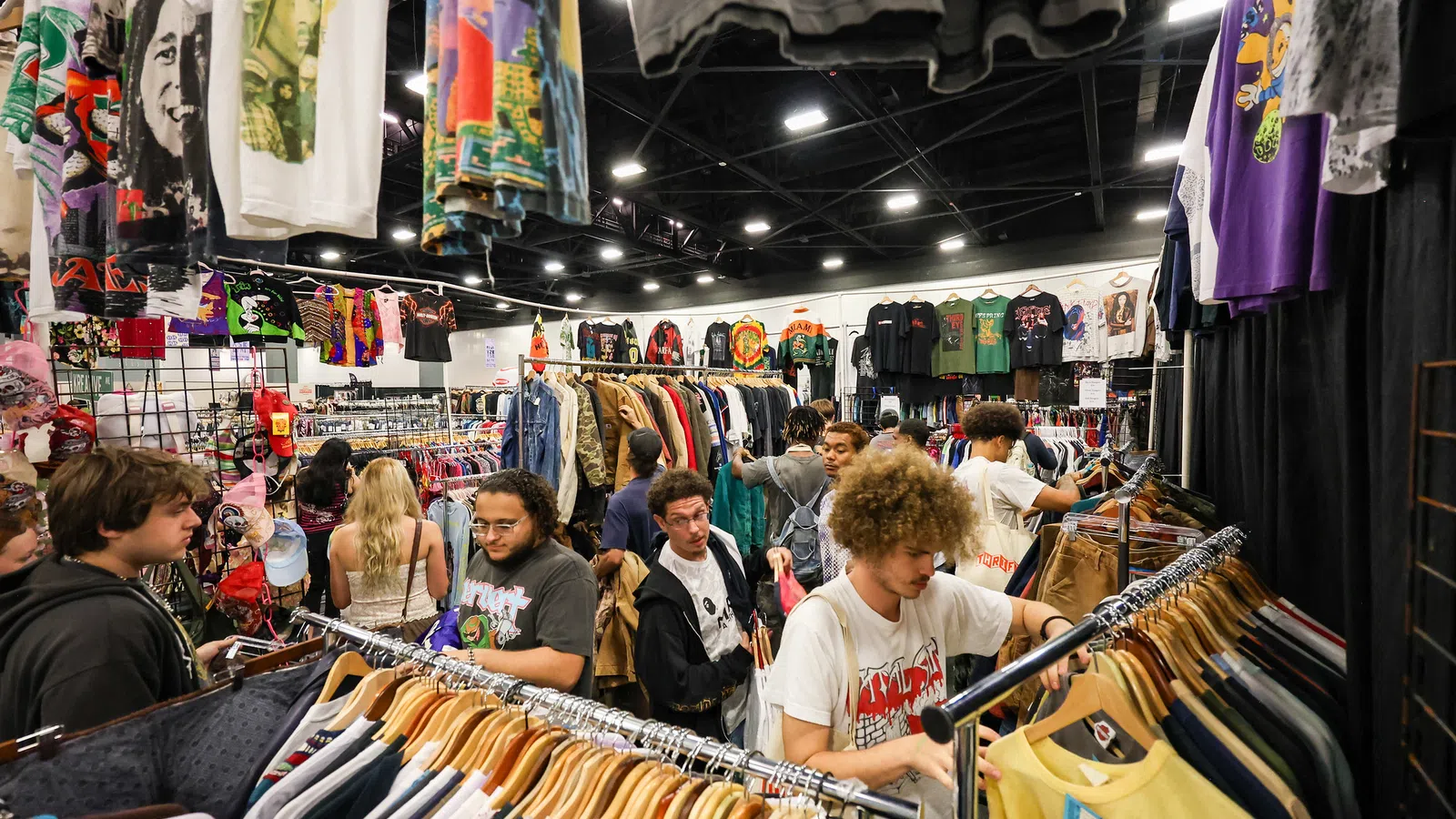 Young shoppers browsing vintage and graphic t-shirts on racks at a busy indoor clothing market.