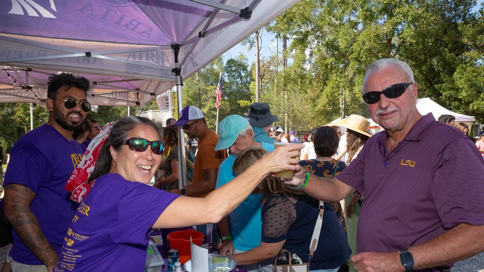 People enjoying a sunny outdoor festival, sharing drinks under a tent with vibrant purple banners.