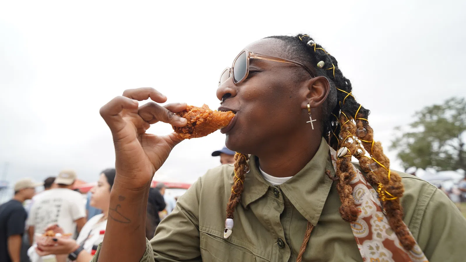 Person with braided hair and sunglasses enjoying crispy fried chicken outdoors at a casual event.