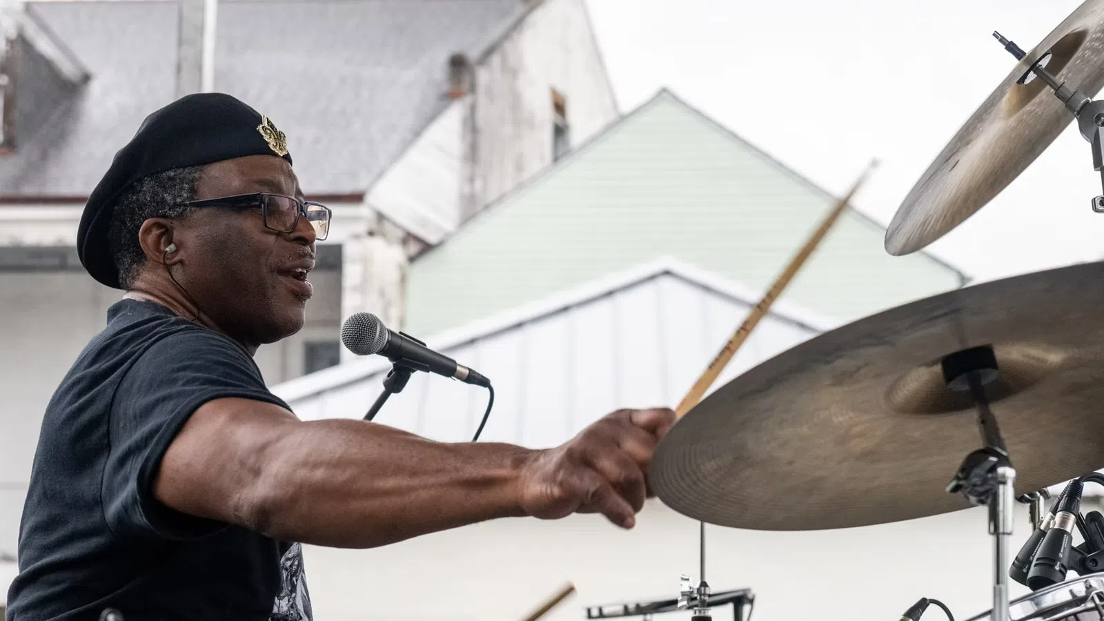 Man wearing a beret playing drums energetically at an outdoor performance with houses in the background