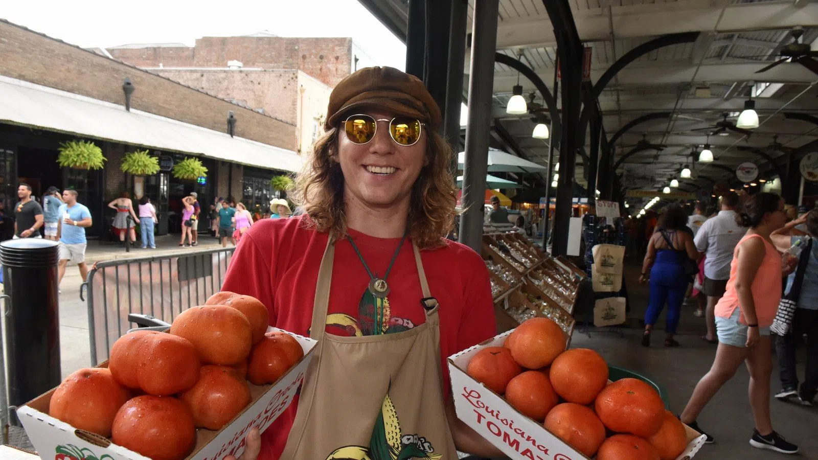 Smiling vendor wearing sunglasses and an apron holds two boxes of fresh Louisiana Creole tomatoes at a busy market.