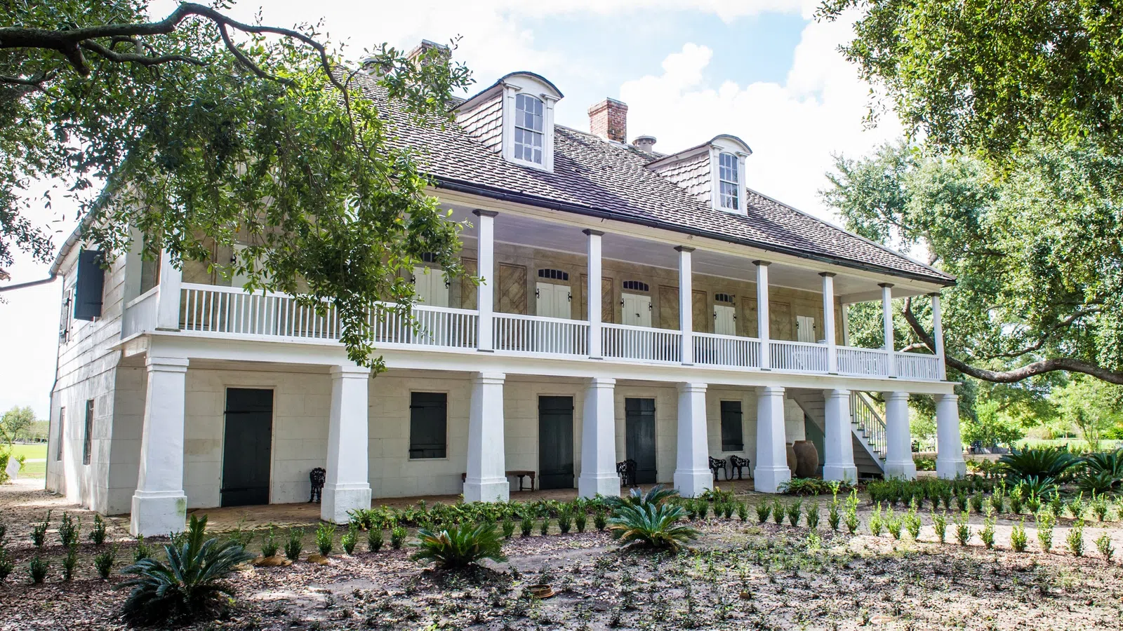 Historic two-story plantation house with white columns, balcony, lush greenery, and a sunny sky background