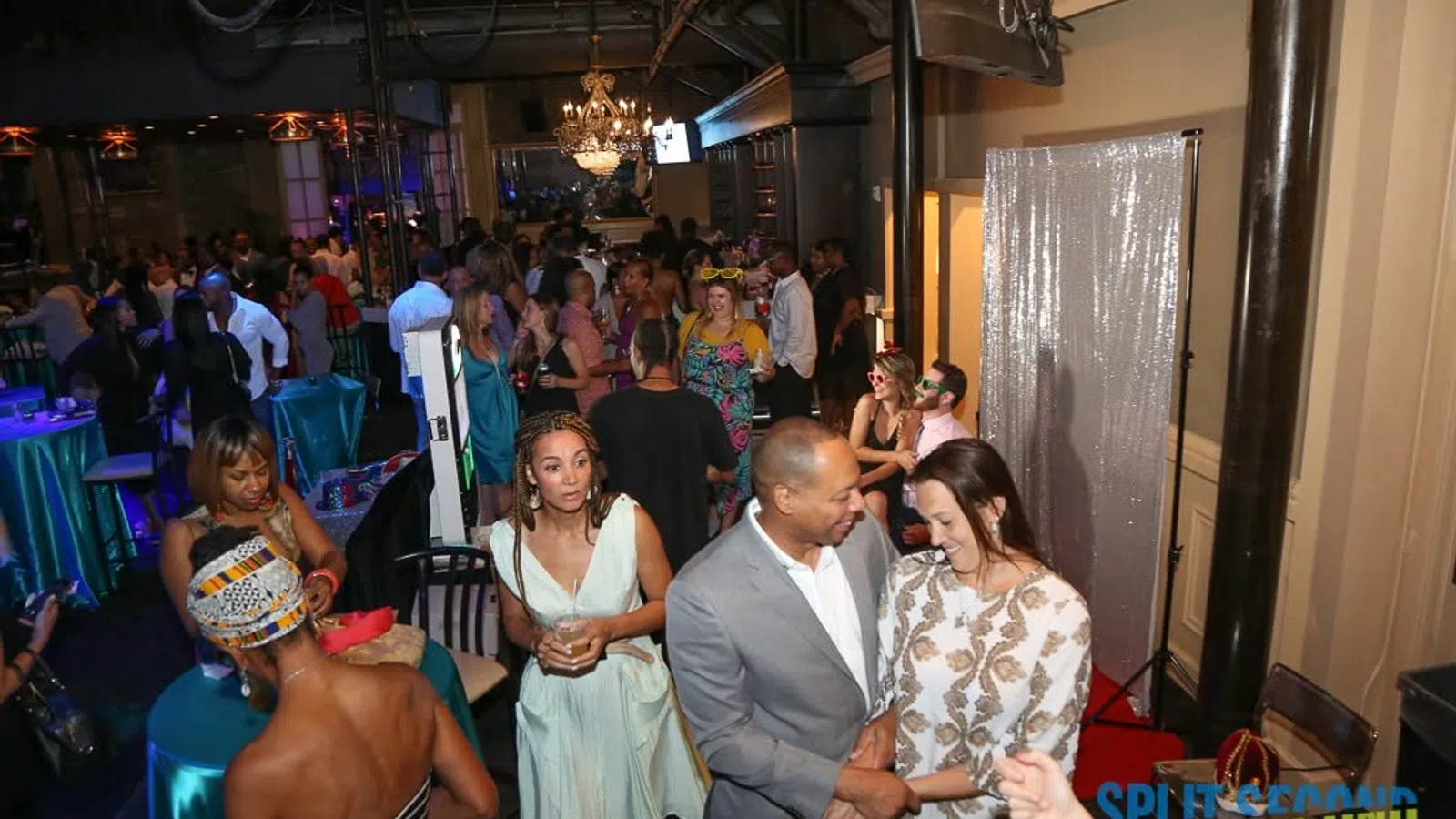 Crowded lively indoor party with guests socializing, dressed in formal and cocktail attire under chandeliers.